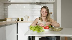 Woman sitting at table preparing fresh vegetables in a modern kitchen, highlighting healthy living and independence.