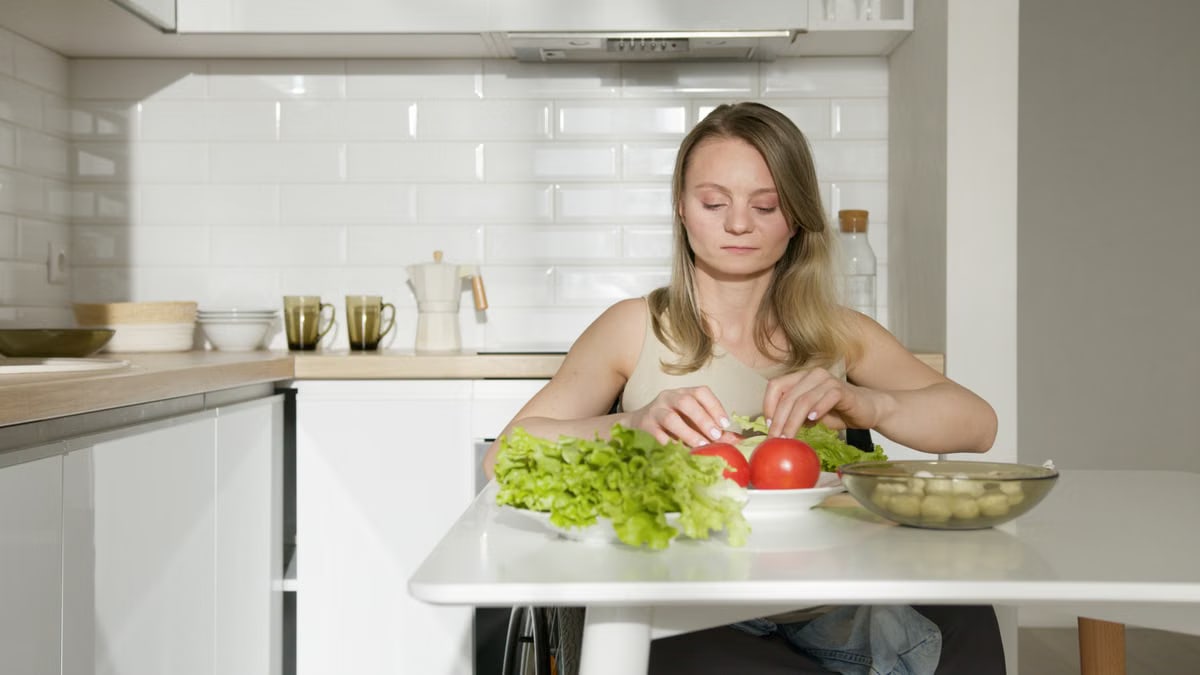 Woman sitting at table preparing fresh vegetables in a modern kitchen, highlighting healthy living and independence.